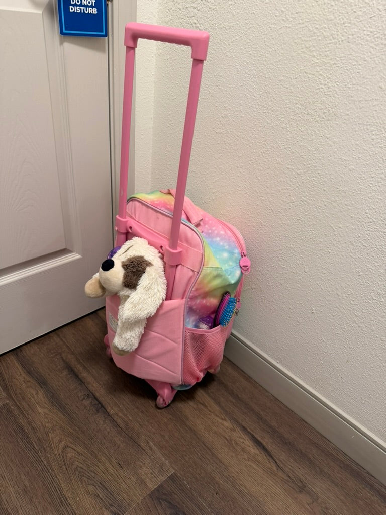 Pink rolling suitcase packed by a child, with a stuffed animal and WetBrush ready for vacation